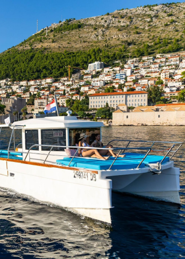 A white boat with people relaxing on the deck sails through blue waters. In the background, a hillside town features colorful buildings and lush greenery under a clear blue sky.