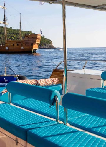 A luxury boat with turquoise cushioned seats overlooks the sea. In the background, a wooden ship sails near a green hilly coastline under a clear blue sky.