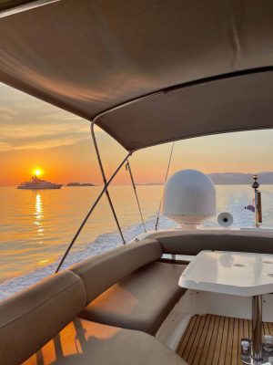 View from a boat at sunset, with a calm sea and an island in the distance. A yacht is silhouetted against the orange sky. The boats deck has cushioned seats and a small table, with a canopy overhead.