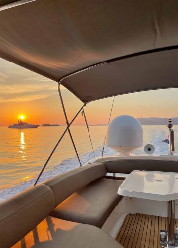 View from a boat at sunset, with a calm sea and an island in the distance. A yacht is silhouetted against the orange sky. The boats deck has cushioned seats and a small table, with a canopy overhead.