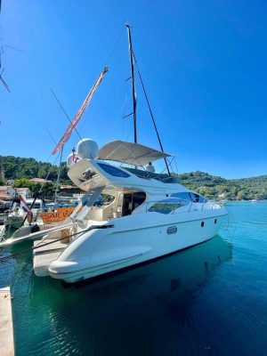 A white yacht is docked in clear blue water under a bright sky. The yacht has multiple decks and is next to a sailboat and a wooded shoreline, suggesting a serene coastal setting.