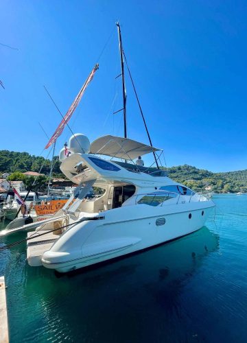 A white yacht is docked in clear blue water under a bright sky. The yacht has multiple decks and is next to a sailboat and a wooded shoreline, suggesting a serene coastal setting.