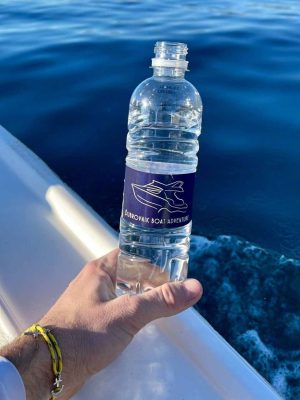 Hand holding a plastic water bottle labeled Dubrovnik Boat Adventure with a boat graphic. The bottle is held over the side of a boat, with blue ocean water in the background and part of the boat railing visible.