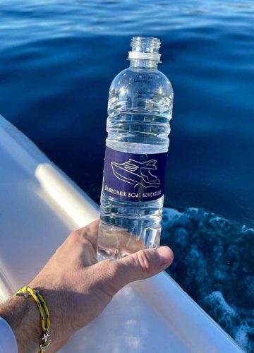 Hand holding a plastic water bottle labeled Dubrovnik Boat Adventure with a boat graphic. The bottle is held over the side of a boat, with blue ocean water in the background and part of the boat railing visible.