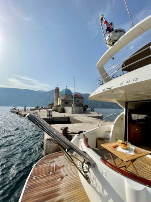 A luxury boat docked near an island with a historic church featuring a blue dome and a bell tower. Calm sea surrounds the island, and a partly sunny sky is above. A wooden deck with fruit on a plate is visible in the foreground.