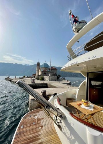 A luxury boat docked near an island with a historic church featuring a blue dome and a bell tower. Calm sea surrounds the island, and a partly sunny sky is above. A wooden deck with fruit on a plate is visible in the foreground.