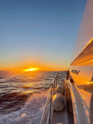 A yacht sails on a calm sea at sunset. The sun is setting on the horizon, casting a warm glow over the waves. The yachts side reflects the golden light, and several fenders line the railing.