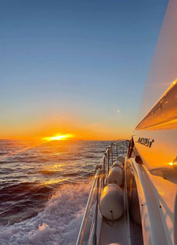 A yacht sails on a calm sea at sunset. The sun is setting on the horizon, casting a warm glow over the waves. The yachts side reflects the golden light, and several fenders line the railing.