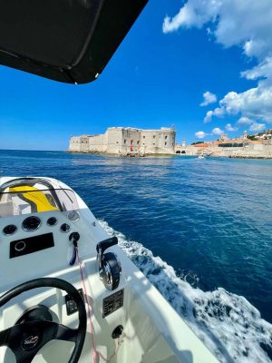 View from a boat approaching a historic stone fortress on the coast. The water is calm, and the sky is bright blue with a few clouds. The boat’s dashboard and controls are visible in the foreground.