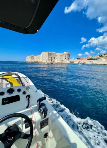 View from a boat approaching a historic stone fortress on the coast. The water is calm, and the sky is bright blue with a few clouds. The boat’s dashboard and controls are visible in the foreground.