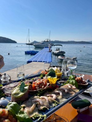 A seafood platter with shrimp, fish, and garnishes on a table by the water. In the background, a blue dock leads to boats moored on a calm sea under a clear sky, with hills in the distance. Glasses and plates are on the table.