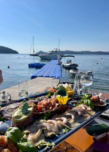 A seafood platter with shrimp, fish, and garnishes on a table by the water. In the background, a blue dock leads to boats moored on a calm sea under a clear sky, with hills in the distance. Glasses and plates are on the table.