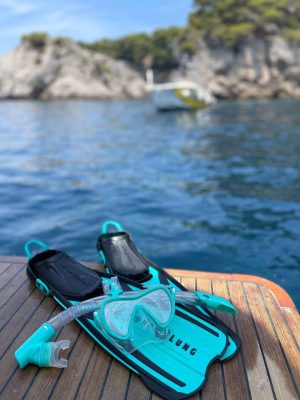 A snorkel set with fins and a mask is placed on a wooden deck of a boat. The turquoise sea and rocky coastline with trees are in the background, along with another boat on the water.
