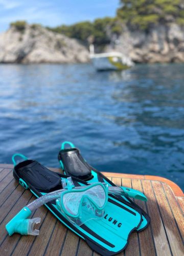 A snorkel set with fins and a mask is placed on a wooden deck of a boat. The turquoise sea and rocky coastline with trees are in the background, along with another boat on the water.