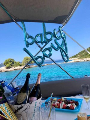 A festive boat setup with blue baby boy balloons displayed. The scene features champagne bottles, a fruit platter, and scenic ocean views with a coastline and trees in the background.