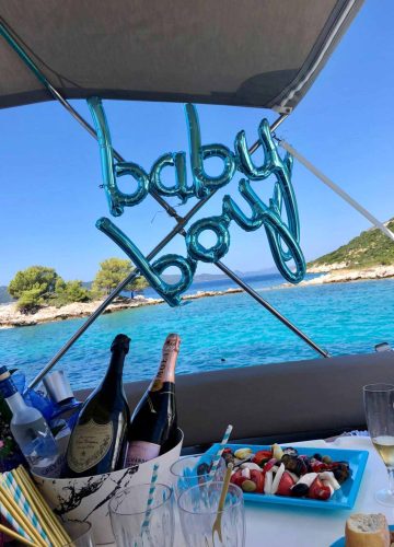 A festive boat setup with blue baby boy balloons displayed. The scene features champagne bottles, a fruit platter, and scenic ocean views with a coastline and trees in the background.