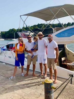 Four adults and a child stand on a dock in front of a white boat. Two men wear white polo shirts and shorts; another man wears a white shirt and hat, holding the child. A woman in a colorful outfit and sunglasses stands to the left. Trees are in the background.