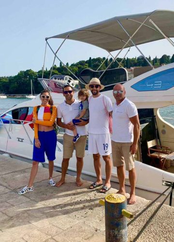 Four adults and a child stand on a dock in front of a white boat. Two men wear white polo shirts and shorts; another man wears a white shirt and hat, holding the child. A woman in a colorful outfit and sunglasses stands to the left. Trees are in the background.