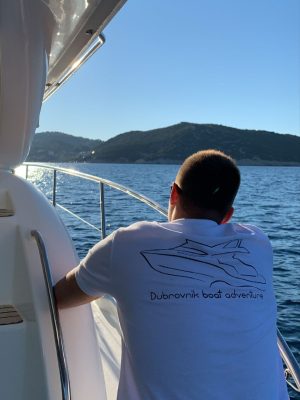 A person wearing a Dubrovnik boat adventure t-shirt leans on the railing of a boat. They gaze at the calm water and distant hills under a clear blue sky.