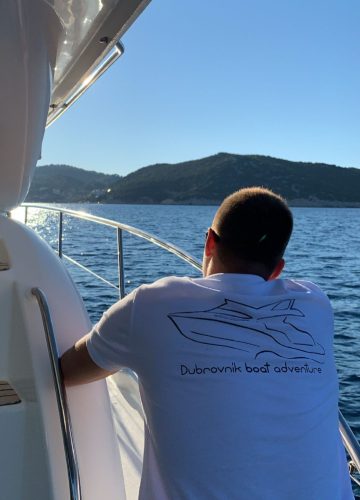 A person wearing a Dubrovnik boat adventure t-shirt leans on the railing of a boat. They gaze at the calm water and distant hills under a clear blue sky.